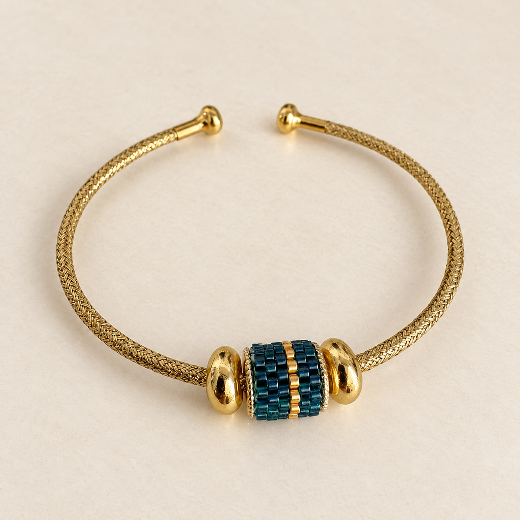 Gold-toned open cuff bracelet with dark teal and gold beaded centrepiece on a warm ivory background