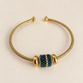 Gold-toned open cuff bracelet with dark teal and gold beaded centrepiece on a warm ivory background
